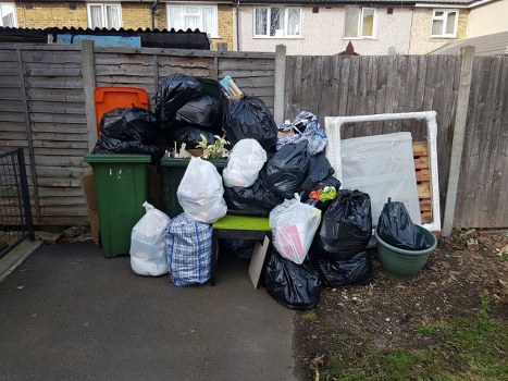 A collection of black, white, and blue striped rubbish bags filled with waste are piled on and around a green plastic bench positioned against a weathered wooden fence in a residential outdoor area. Behind the pile, there is a larger green wheelie bin with a closed lid and an orange recycling box, also placed against the fence. To the right, a white metal bed frame with no mattress is leaning upright, partially obscured by the rubbish bags. A large, round, dark-green plastic container or planter is situated near the bed frame on the dirt ground. The scene takes place on a paved driveway, with a grey concrete surface in the foreground, and the fence encloses the garden or yard area. In the background, the upper floors of nearby houses with light-colored walls and multiple windows are visible, indicating a typical residential setting possibly in or near [TOWN] or [POSTCODE]. The overall scene depicts waste accumulation typical of garden or household clearance, and it reflects the type of rubbish removal service offered by Garden Clearance Temple in the local area.