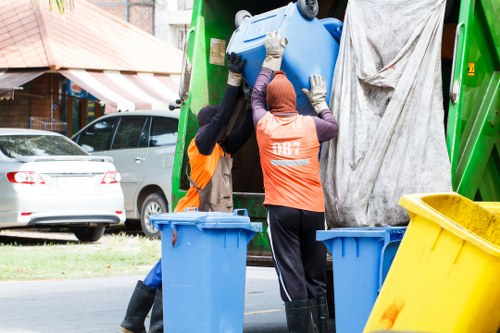 Two waste collection workers wearing high-visibility orange vests and protective gloves are handling a large blue plastic rubbish bin, lifting it towards a green waste collection vehicle parked on a residential street in the vicinity of the postcode area. One worker is partially obscured, while the other, with their back to the camera, is guiding the bin into the vehicle. The waste vehicle has a grey fabric cover partially draped over its rear, and the surrounding environment includes a grey car parked on the pavement, a house with a brown roof and a partially visible awning, and a grassy verge. The scene is set in natural daylight, indicating daytime conditions typical for rubbish removal in the local area, and the image visually supports professional waste collection services such as those offered by Garden Clearance Temple across [TOWN] or nearby postcode districts.