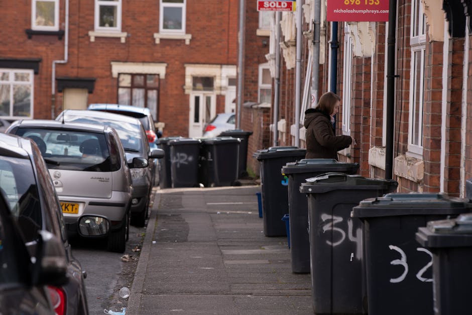 A middle-aged woman with dark hair tied back is seen cleaning a public outdoor area by disposing of rubbish into a tall, stainless steel litter bin. She is wearing a black T-shirt with printed white t