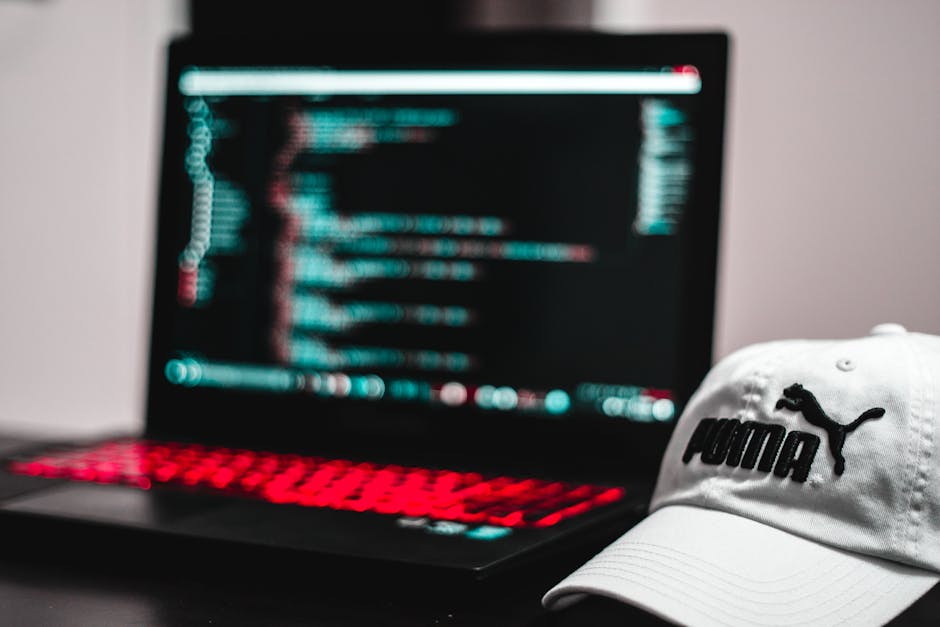 A black laptop computer displaying lines of multicoloured code on its screen, positioned on a flat surface with a slight tilt showing a close-up view. The laptop has a partially visible red backlit ke