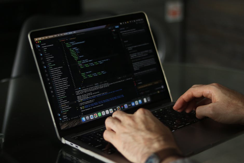 A person with short dark hair, dressed in a light blue shirt, is sitting at a workspace and working on a laptop computer that displays lines of multicolored programming code on its screen. The desk fe